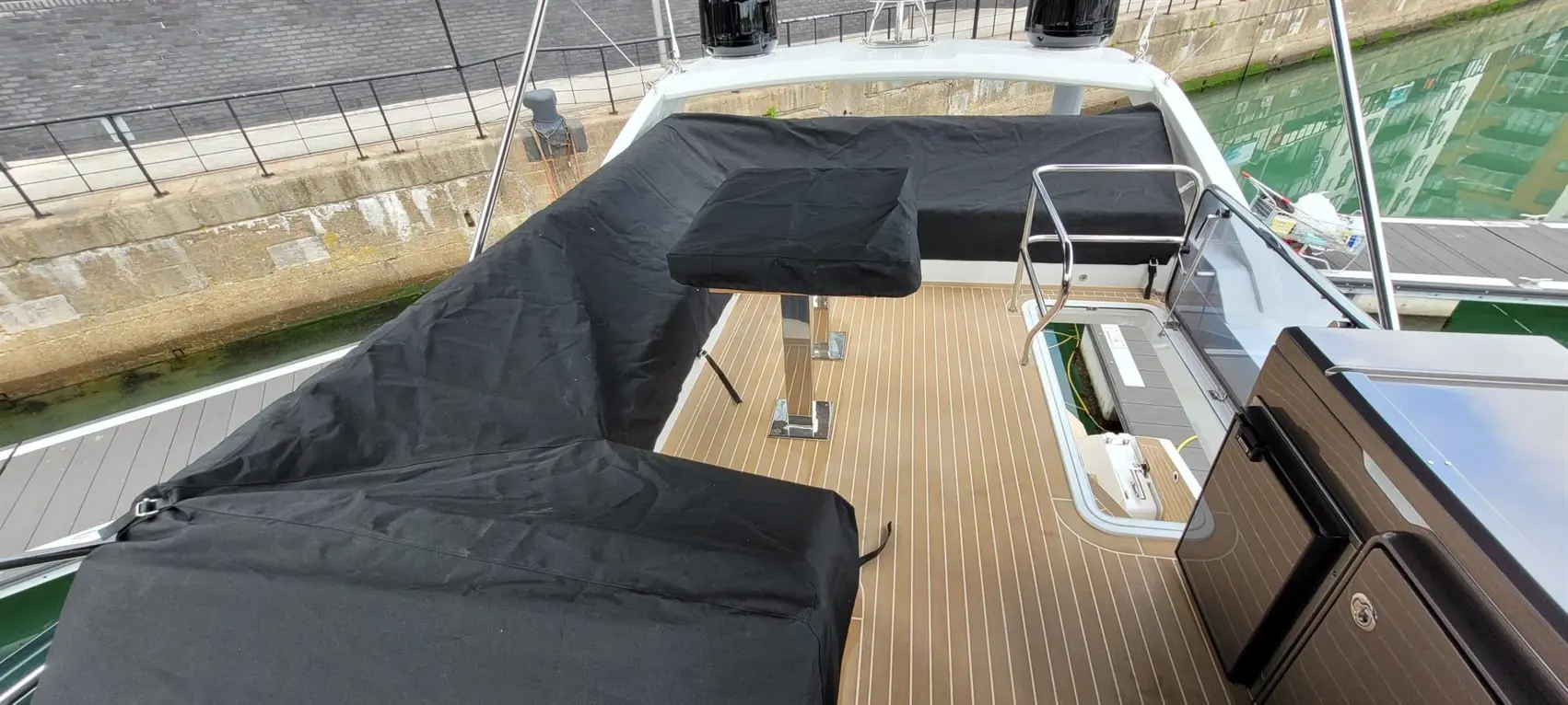 Overview of a boat deck with black covers over seating, stainless steel table leg, and a glimpse of the wharf nearby.